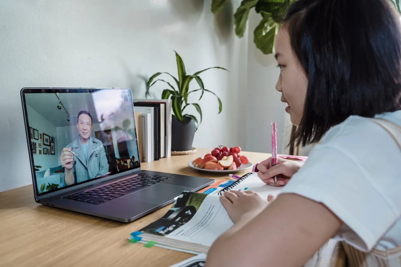 Pessoa acompanhando uma videochamada no notebook e anotando em um caderno, com o material de estudo aberto sobre a mesa